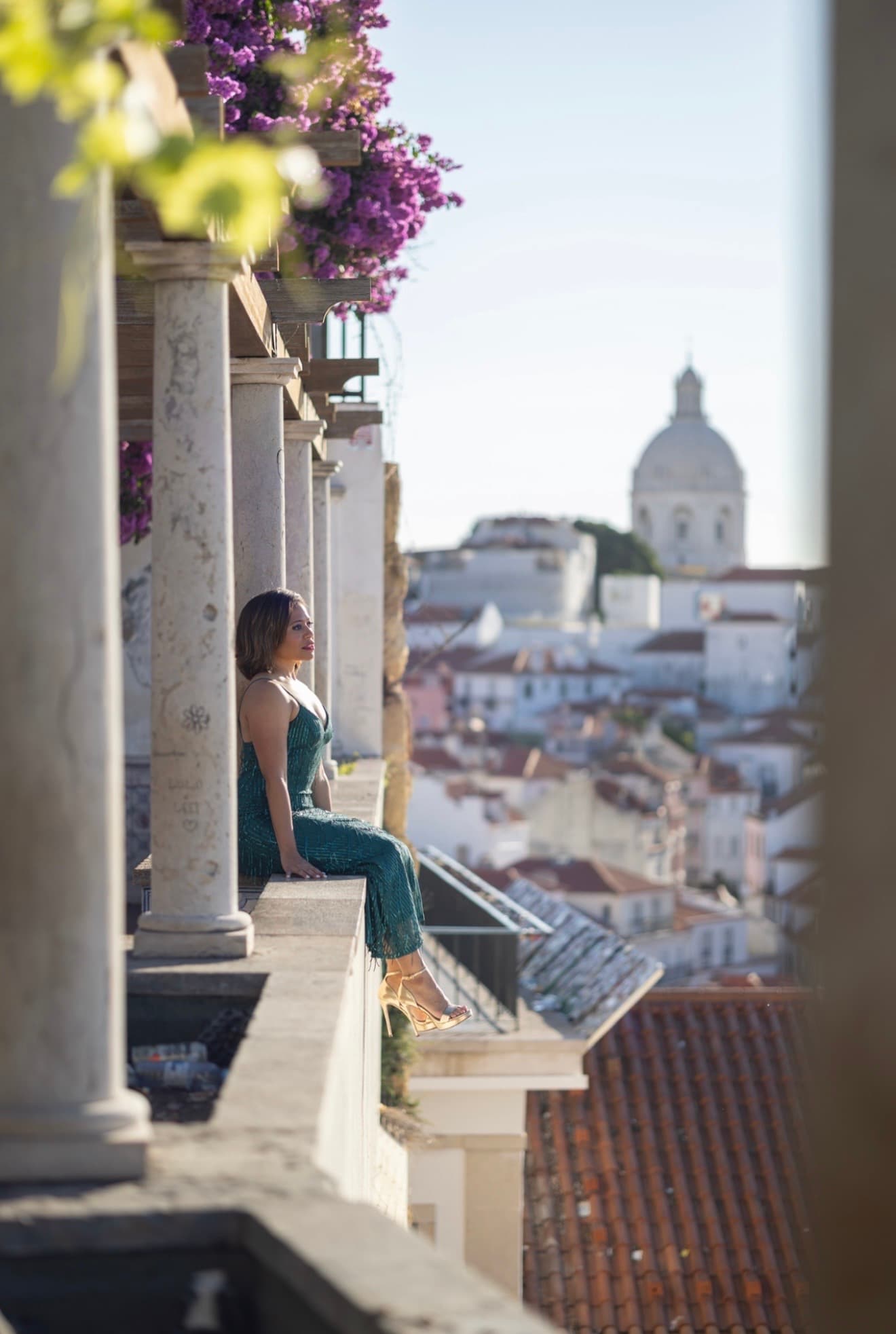 Woman in a green dress sits on a balcony overlooking a sunlit European cityscape.