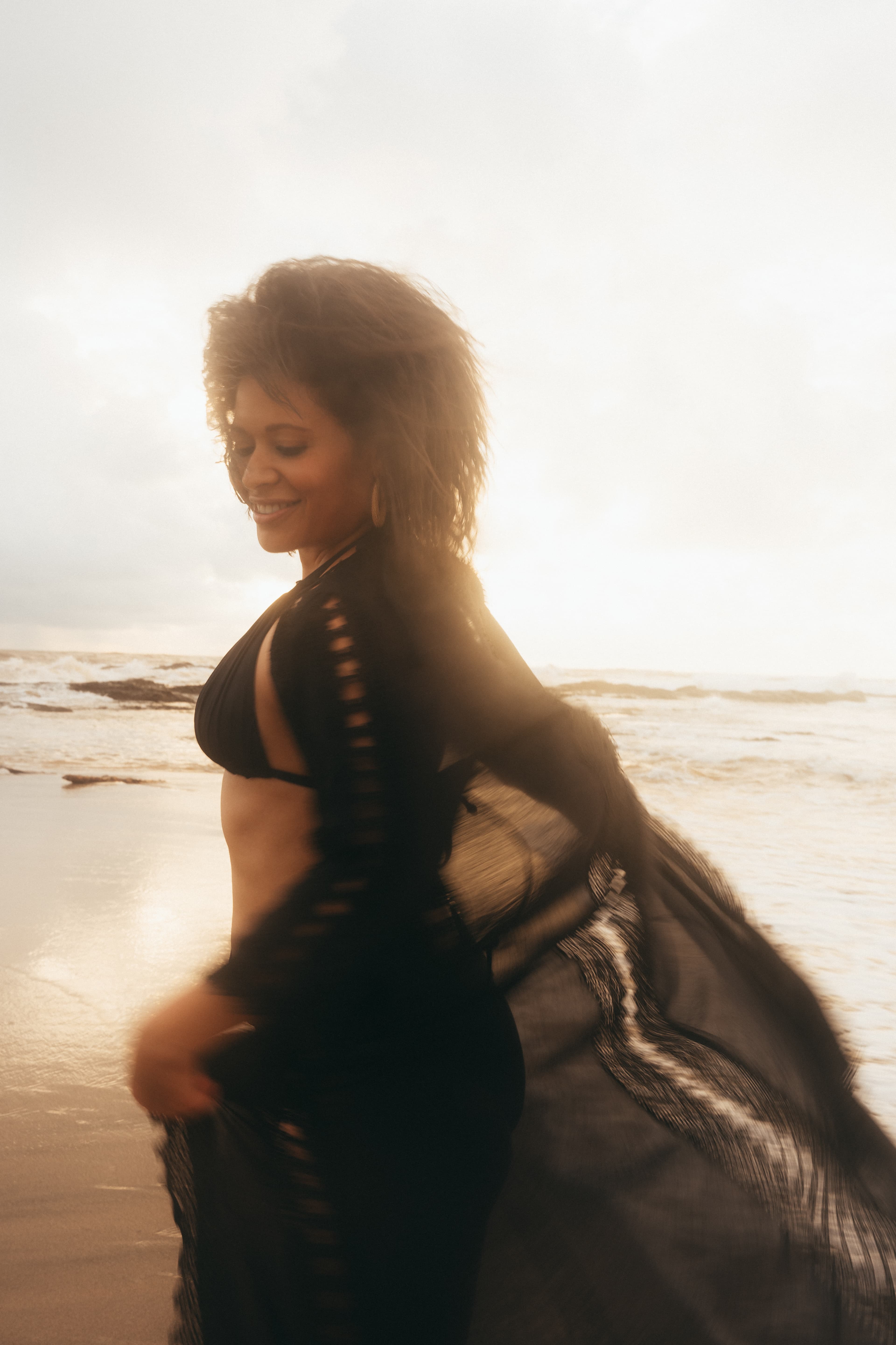 Smiling woman in a black bikini and flowing cover-up on a beach during golden hour sunset.