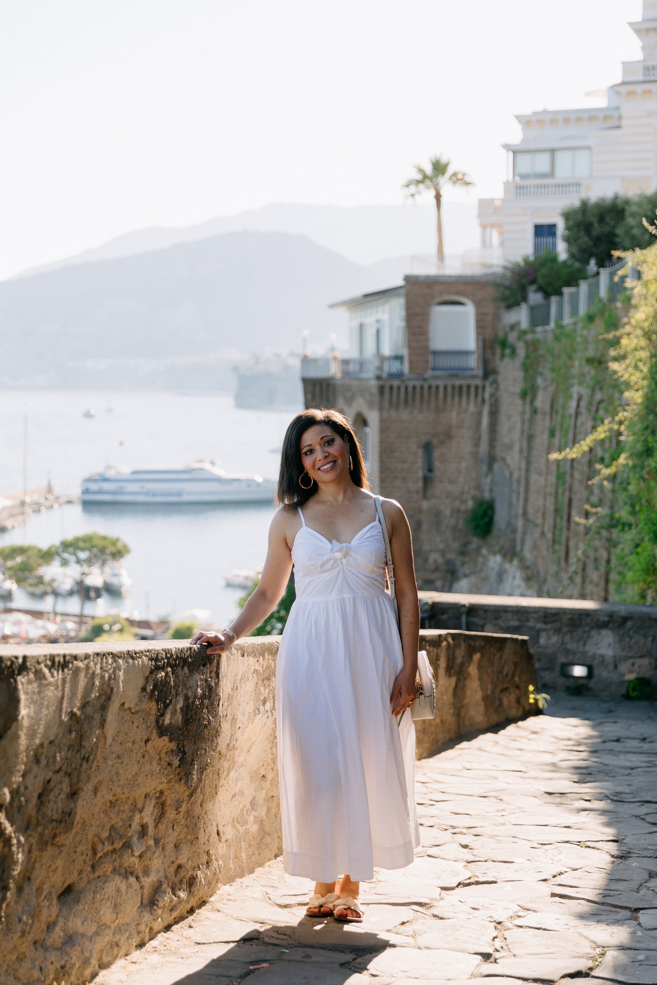 Smiling woman in a white dress stands on a stone path overlooking a coastal harbor.